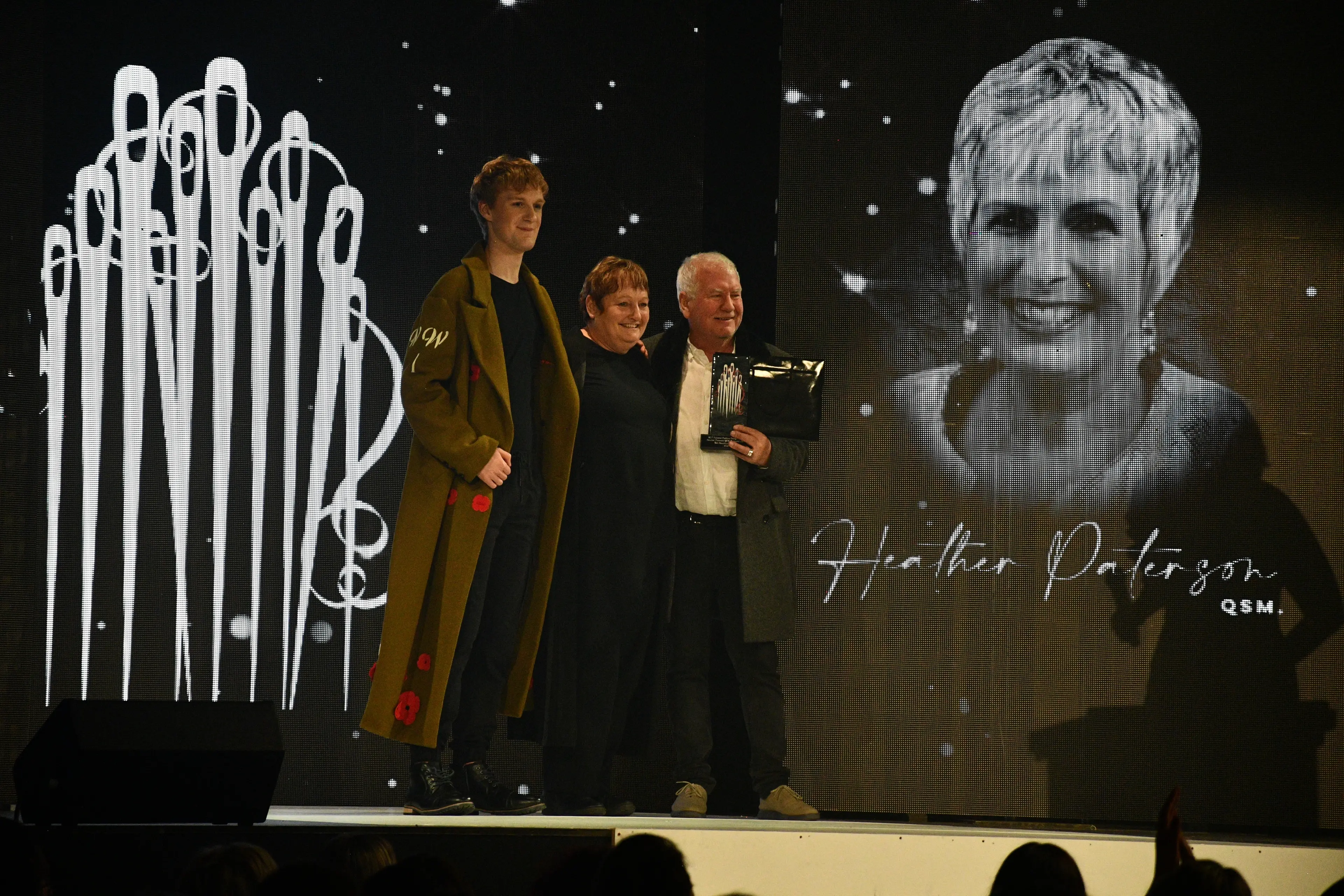 Dorothy McIntosh (centre) wins the Heather Paterson QSM Memorial Award for Best Southland Designer for her Anzac-themed coat, at the Hokonui Fashion Design Awards on Saturday. The award is presented to her by Wade Paterson, (right) and the garment is worn by model Jude Terry. Photo: Gemma Sinclair