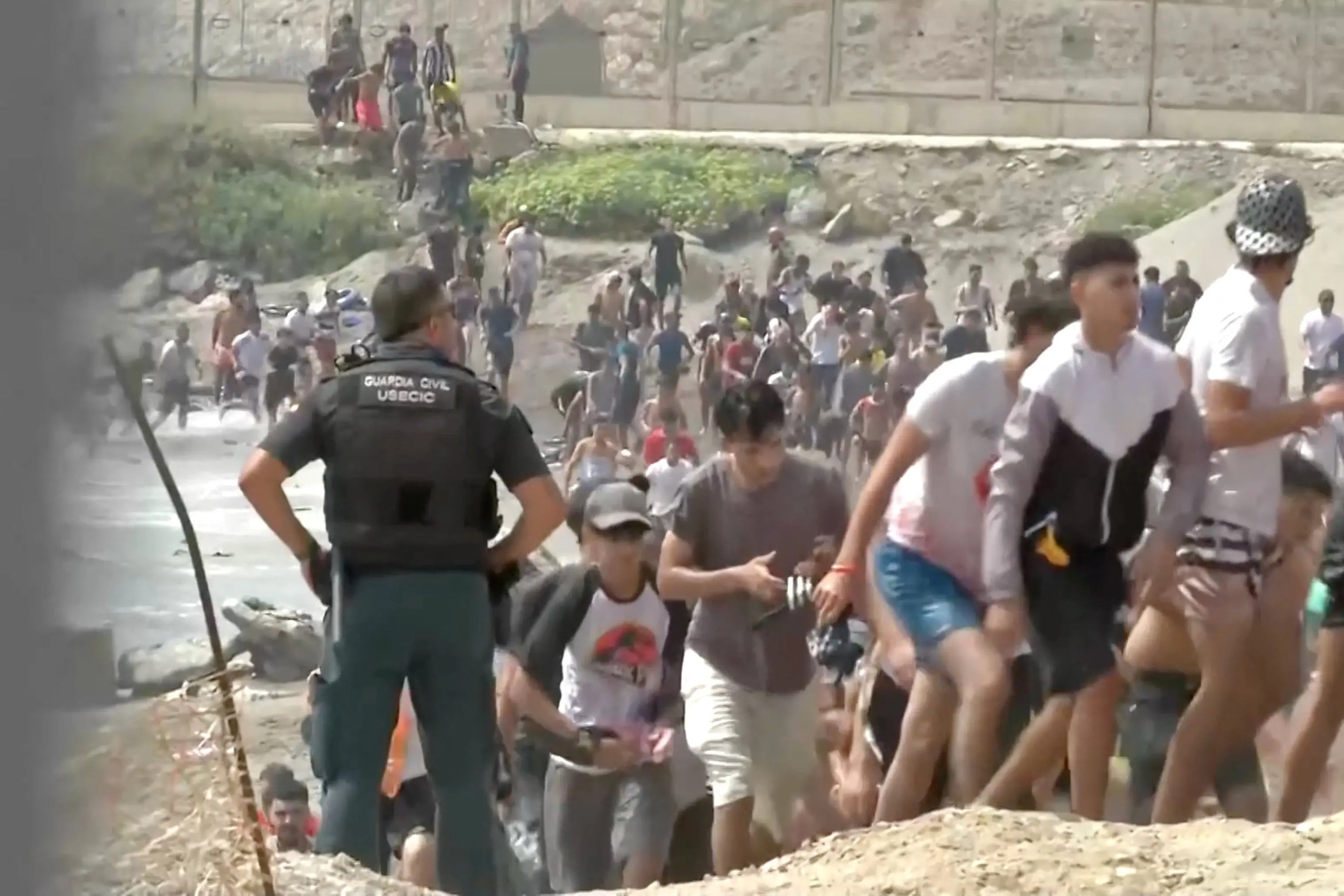 <p>Migrants walk next to a USECIC Guardia Civil member after swimming through Spain's Ceuta border from the Moroccan side to reach the border fence, in Ceuta, Spain. Photo:&nbsp;Atlas/via Reuters TV/Handout via REUTERS</p>\\n